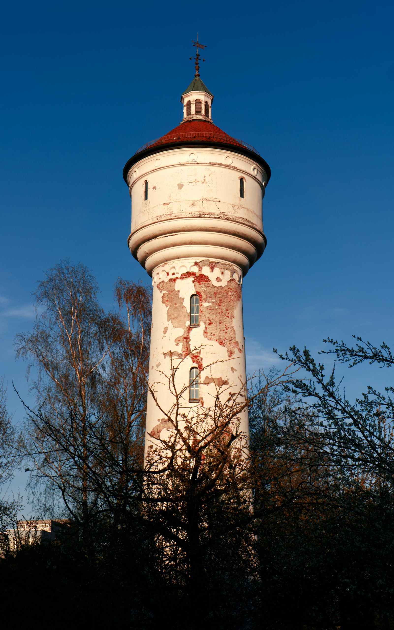 White tower with red coned roof against a blue sky with bushy trees in the foreground.